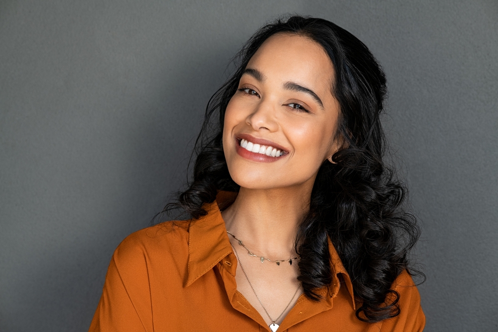 Smiling young woman with long, dark curly hair, wearing an orange shirt and layered necklaces, posing in front of a plain gray background.